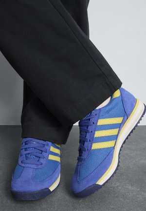 Blauwe sport schoenen met gele strepen, gemaakt van mesh en suedemateriaal, ronde neus, textuur op het oppervlak en een witte tussenzool, gecombineerd met zwarte broek.