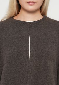 Donna che indossa un cardigan di lana marrone scuro con scollatura rotonda, fissato da un singolo bottone nella parte superiore, sopra un top di colore chiaro.