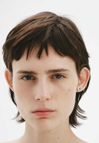 Cabello corto y oscuro con mechones despeinados, ojos azules y lleva pequeños pendientes de plata redondeados con una piedra azul clara. Fondo sencillo.