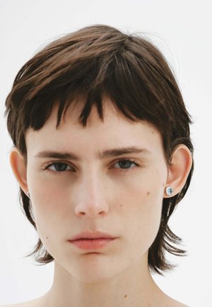 Cabello corto y oscuro con mechones despeinados, ojos azules y lleva pequeños pendientes de plata redondeados con una piedra azul clara. Fondo sencillo.