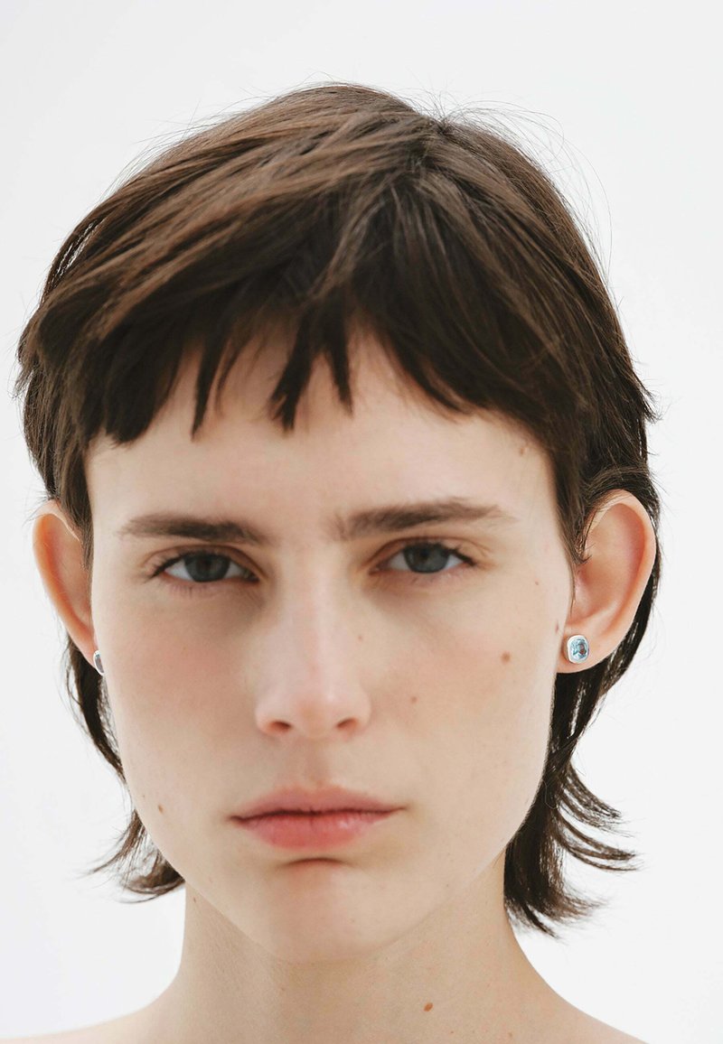 Cabello corto y oscuro con mechones despeinados, ojos azules y lleva pequeños pendientes de plata redondeados con una piedra azul clara. Fondo sencillo.