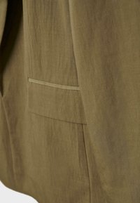 Gros plan sur la poche d'une veste en tissu kaki avec un détail de bordure horizontale et une couture visible sur un matériau texturé.