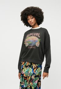 Sweatshirt noir avec un motif "RIPCURL HAWAII", coupe décontractée, encolure côtelée, porté avec un pantalon multicolore à motif floral.