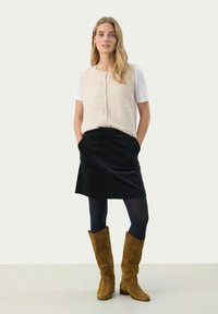 Beige gebreide vest met knopen over een witte t-shirt, gecombineerd met een zwarte corduroy rok en bruine suede laarzen tot aan de knie.