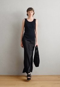 Modella che indossa un top senza maniche a coste nero e una gonna maxi in satin nero, con in mano una borsa plissettata nera, su un pavimento in legno chiaro contro un muro grigio.