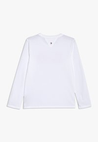 Biała bluzka z długim rękawem, okrągłym dekoltem i małą czerwoną gwiazdką w trójkątnym wycięciu na plecach. Wykonana z miękkiej bawełny, o podstawowym designie.