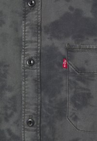 Szara koszula z denimu z delikatnym marmurkowym wzorem, czarnymi guzikami oraz małą czerwoną metką z logo przy kieszeni na piersi.