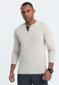 Mann, der ein beiges Langarm-Henley-Shirt mit Holzknöpfen und eine schwarze Armbanduhr trägt, steht vor einem einfarbigen hellen Hintergrund.