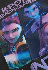 Fekete póló, melyen három animált karakter fényes nyomtatással szerepel, mindegyik egyedi frizurával és színes ruházattal, felettük a "KPOP DEMON HUNTER" felirat.