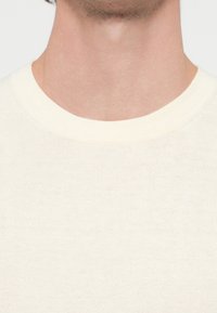 Kremowy sweter dziany z okrągłym dekoltem i gładką fakturą, ukazujący subtelne odcienie przędzy i brak widocznych wzorów.