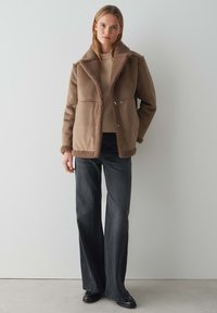 Bruine schapenvacht jas met een brede kraag en een zachte, gestructureerde voering. Voorzien van een knoopsluiting en gecombineerd met donkere flared jeans.