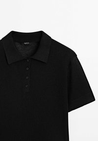 Musta neulepolo, jossa on ribattu kaulus ja lyhyet hihat. Tässä paidassa on neljä mustaa nappia ja teksturoitu kangas.