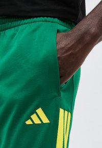 Χέρι με σκούρο δέρμα στην τσέπη πράσινου αθλητικού σορτς με κίτρινες ρίγες και κίτρινο λογότυπο Adidas, φορώντας μαύρο μπλουζάκι.