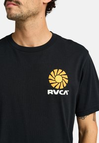 Sort bomulds T-shirt med et gult solgrafik og hvid "RVCA" logo på venstre bryst. Korte ærmer og rund halsudskæring.