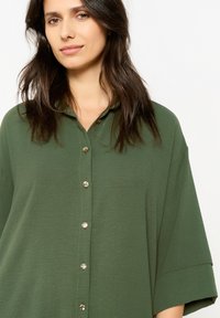Green button-up shirt with a collar, short sleeves, and a relaxed fit; features metallic buttons and a soft, textured fabric.