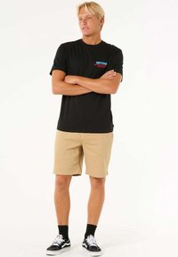 Sort bomulds t-shirt med farvet logo, beige shorts og sorte sneakers med hvide detaljer. Modellen står med armene i kors.