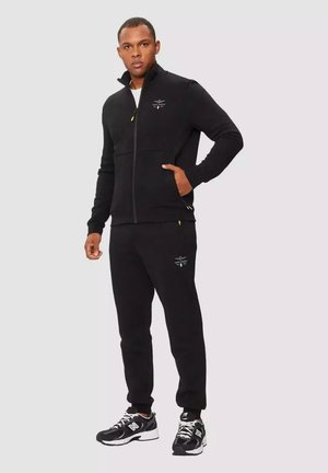 Giacca nera con zip e colletto alto, con logo ricamato e dettagli gialli; abbinata a pantaloni jogger neri e sneakers nere.