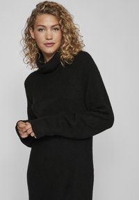 Maglione a collo alto nero realizzato in un morbido e spesso tessuto a maglia, caratterizzato da maniche lunghe e polsini a costine, che arriva sotto il ginocchio per un fit rilassato.