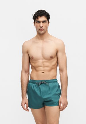 Ung mand med mørkt hår, uden skjorte, iført teal badebukser med en sort-hvid snor, stående mod en ensfarvet lys baggrund.