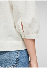 Blusa bianca in lino con maniche 3/4, caratterizzata da polsini arrotolati con bottoni e una sottile trama. Abbinata a jeans in denim blu.