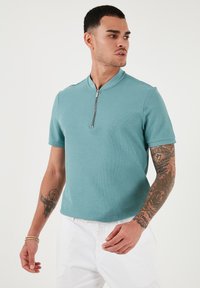 Polo majica s kratkimi rokavi v teal barvi z zadrgo pri ovratniku. Narejena iz teksturirane tkanine, ima sodoben kroj in čist, minimalističen dizajn.
