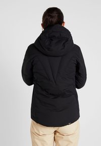 Veste matelassée noire avec capuche ajustable, présentant une texture lisse et un design matelassé, s'arrêtant à la taille, associée à un pantalon de couleur claire.