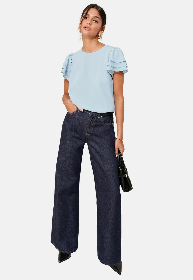Donna che indossa una blouse con maniche a volant di colore azzurro chiaro, jeans a gamba larga scuri, scarpe nere con punta, mentre tiene una piccola borsa nera su uno sfondo bianco.