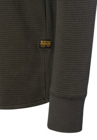 Ciemnozielona, fakturowana koszula z długim rękawem, z prążkowanym mankietem oraz małą metką G-Star w kolorze żółtym i czarnym na rękawie.