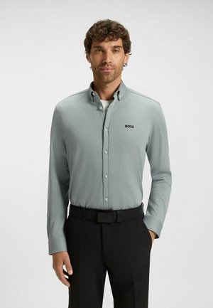 Man in een lichtgrijze lange mouwen overhemd met 'BOSS' logo, zwarte riem en zwarte broek, staande tegen een effen achtergrond.