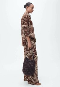Brown patterned outfit featuring paisley designs, paired with a textured black woven bag. Model stands in profile against a plain background.