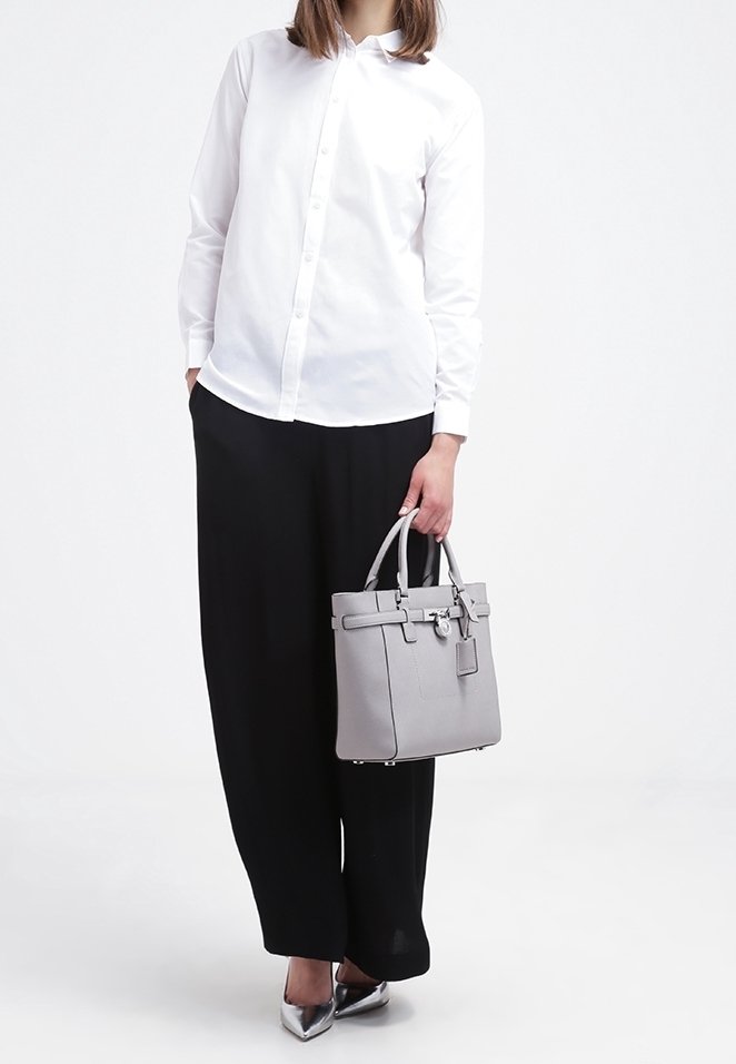 Sac à main en cuir gris avec deux poignées, de forme rectangulaire et avec des accessoires en argent. Le mannequin porte une chemise blanche à boutons et un pantalon large noir.