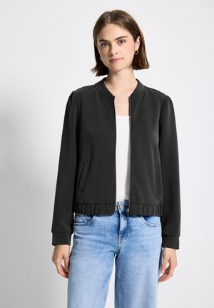 Veste bomber noire avec un ourlet côtelé, fermeture éclair, poches latérales et manches longues, portée sur un haut blanc et associée à un jean bleu clair.