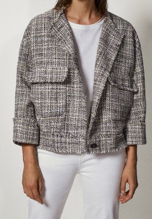 Personne portant une veste courte en tweed texturé sur une chemise blanche et un pantalon blanc, debout devant un fond uni.
