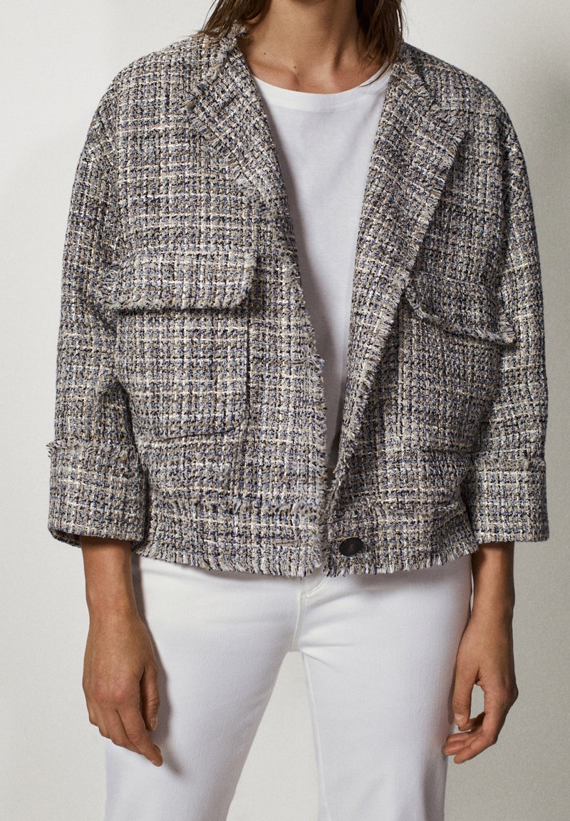 Personne portant une veste courte en tweed texturé sur une chemise blanche et un pantalon blanc, debout devant un fond uni.