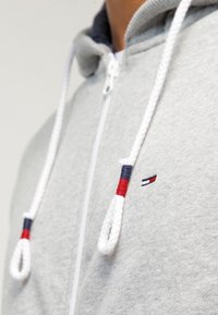 Ljusgrå zip-up hoodies med en mörknavy-fodrad huva, med vita dragsnören med röda och blå detaljer, samt en liten broderad logotyp på bröstet.