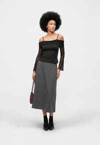 O femeie cu coafură afro purtând un top negru cu umerii goi și mâneci transparente, o fustă midi gri, șosete negre și pantofi negri, ținând o geantă de mână mov.