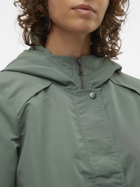 Nahaufnahme einer Person, die eine grüne Kapuzenjacke mit Reißverschluss und geknöpftem Überzug trägt, mit lockigem Haar und großen Creolen.
