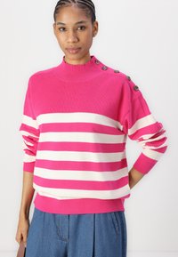 Pull rayé rose et blanc avec un col montant, texture côtelée et accents de boutons sur l'épaule. Associé à un pantalon plissé bleu.