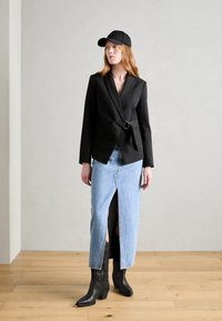 Svart blazer med belte i livet, lys blå denimskjørt med frontsplitt, og svarte cowboystøvler. Modellen har på seg en svart caps.