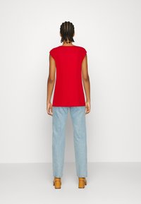 Henkilö, jolla on letitetyt hiukset, pukeutuneena hihattomaan punaiseen paitaan, vaaleansiniset farkkuihin ja ruskeisiin korkokenkiin, seisoo selkä kohti kuvaa valkoista taustaa vasten.