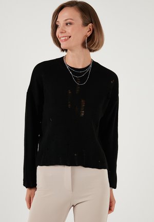 Pulover negru din tricot cu detalii decupate, guler rotund și mâneci lungi, asortat cu pantaloni ajustați de culoare crem.