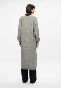 Vestido de suéter largo gris de punto con un ajuste relajado, hombros caídos y dobladillo acanalado, combinado con pantalones negros de pierna ancha.