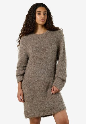 Femme aux longs cheveux bouclés, portant une robe-pull ample et texturée couleur taupe, debout sur un fond blanc uni.