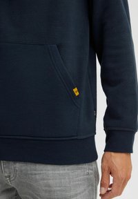Navy fleece sweatshirt with a front pocket, ribbed cuffs, and a small yellow label on the hem, displayed alongside gray pants.