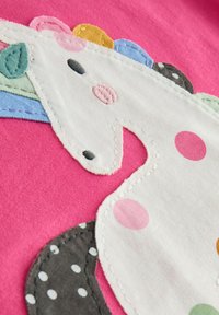 Mehrfarbige Einhornapplikation auf einem rosa Stoffhintergrund, verziert mit weißen Tupfen sowie gestickten Details in unterschiedlichen Farben und Texturen.