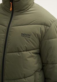 Olivengrøn isoleret dunjakke med høj krave, sort lynlås og broderet logo. Har quiltet syning og orange lynlåstræk.