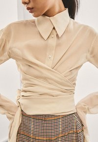 Chemise beige à boutons avec un design croisé, manches longues et un col, associée à une jupe à carreaux aux motifs marron et orange.