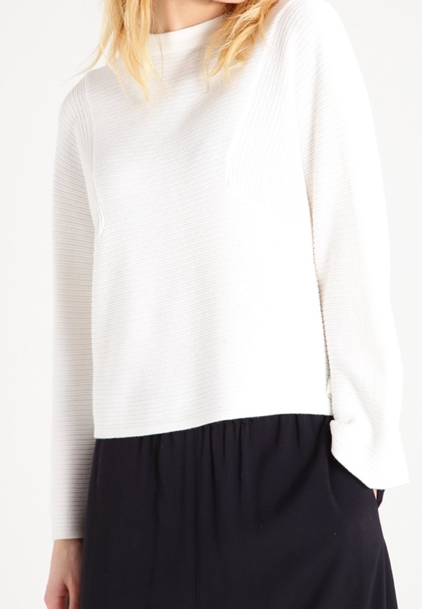 Vit ribbad tröja med långa ärmar och rund halsringning, tillsammans med en svart kjol. Den texturerade tyget ger ett subtilt visuellt intresse.