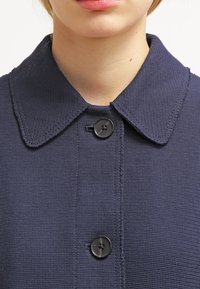 Námořnická modrá strukturovaná bunda s límcem, která má vpředu dvě černé knoflíky. Tkanina vypadá jako tkaná a je pevná.