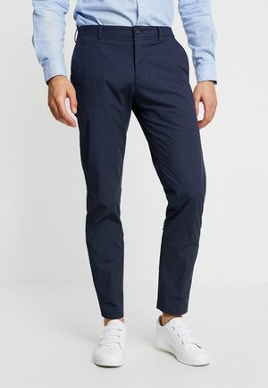 Man draagt slanke donkerblauwe broek, lichtblauw overhemd en witte klittenbandsneakers, staand tegen een effen achtergrond.